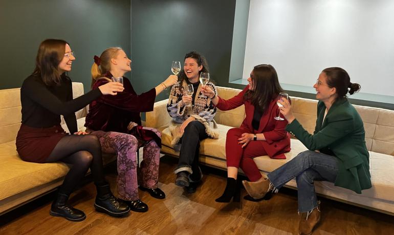 Five women toasting together on a beige couch.